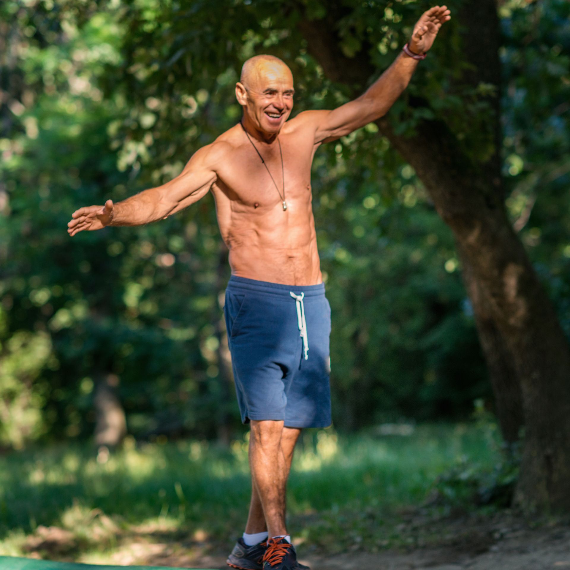 A shirtless man in blue shorts is standing in the woods with his arms outstretched.