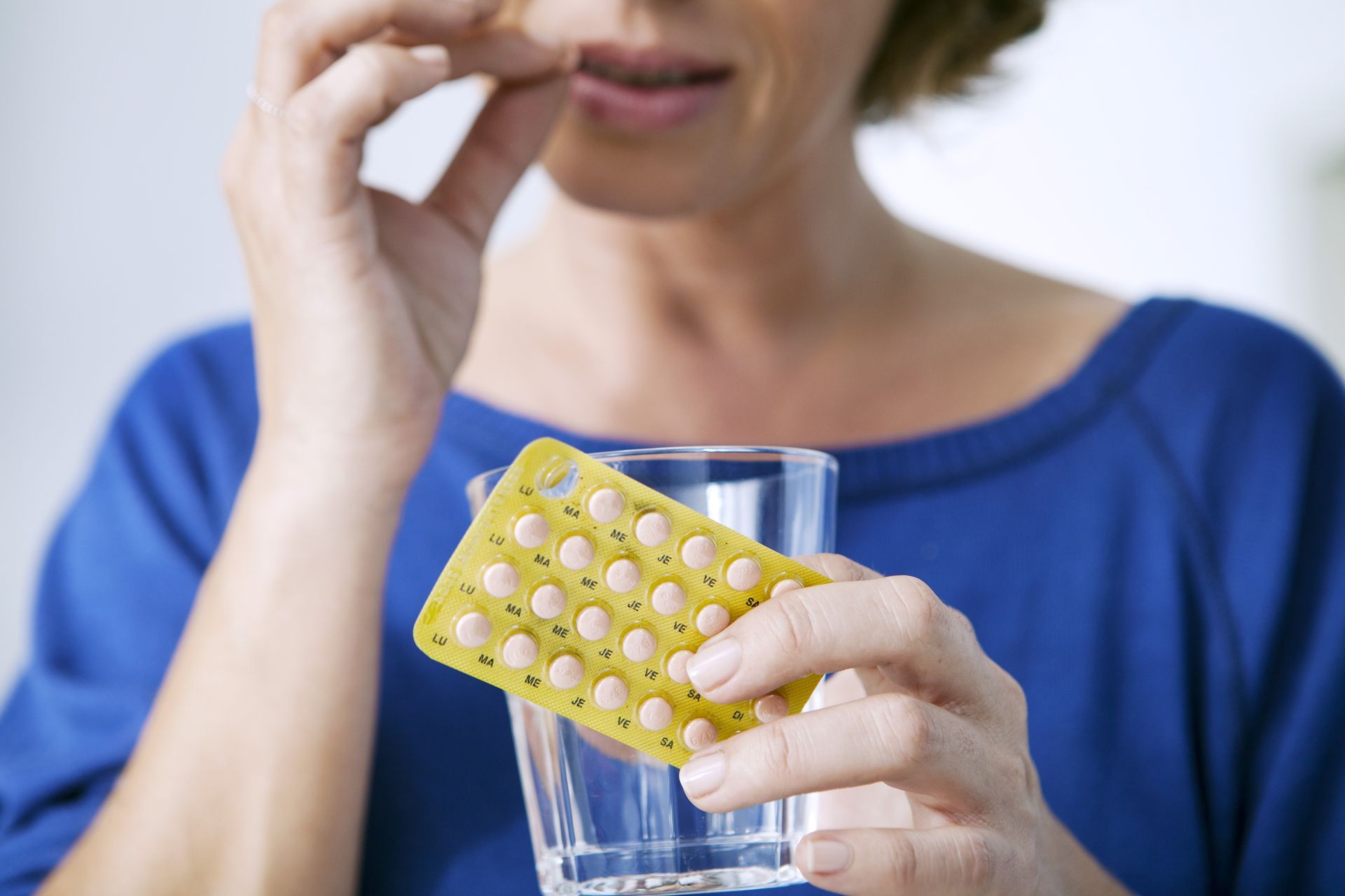 A woman is taking a pill from a blister pack while holding a glass of water.