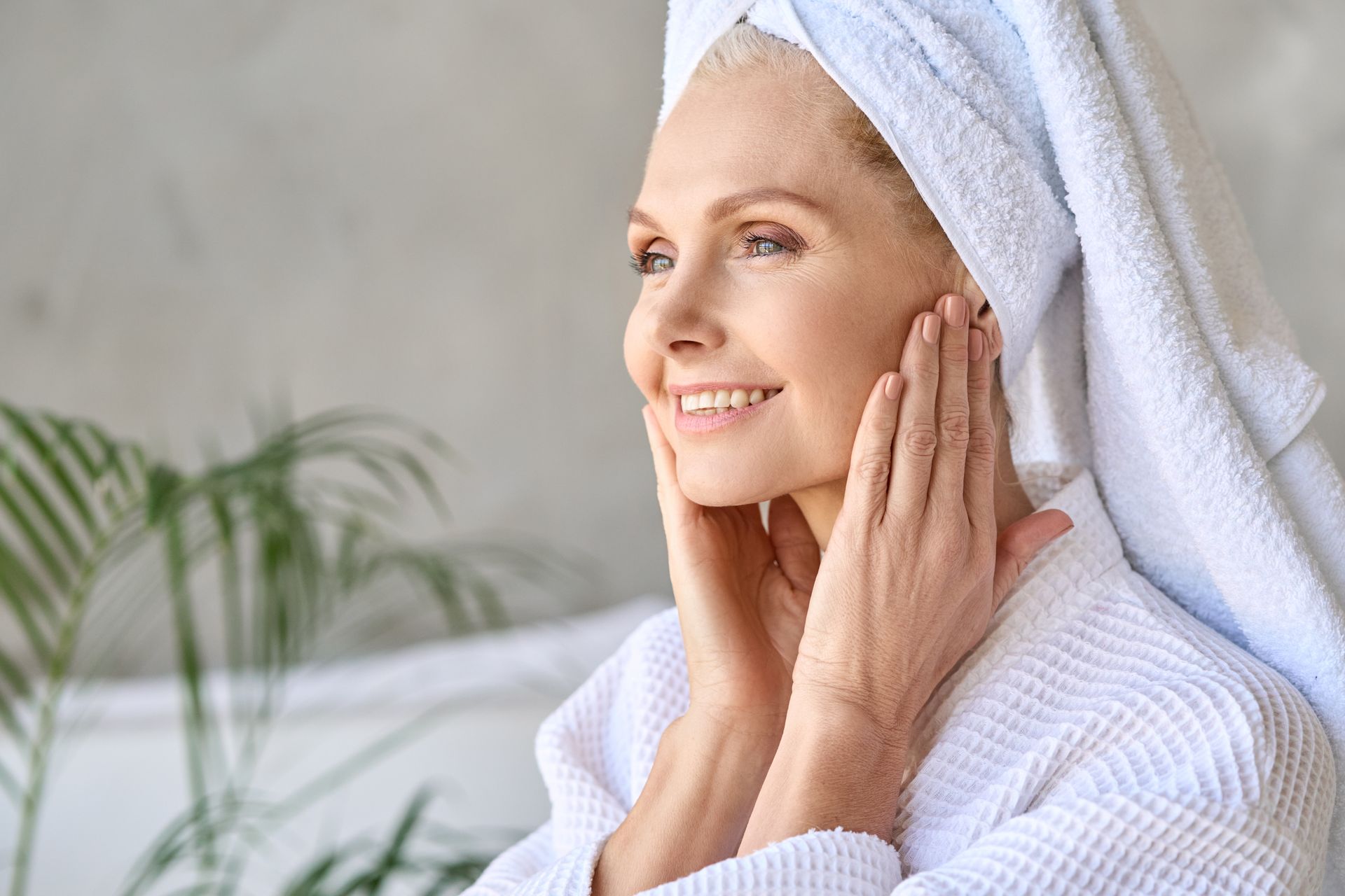 Woman in white robe and towel, hands on face, smiling, with a background of a plant and bathroom.