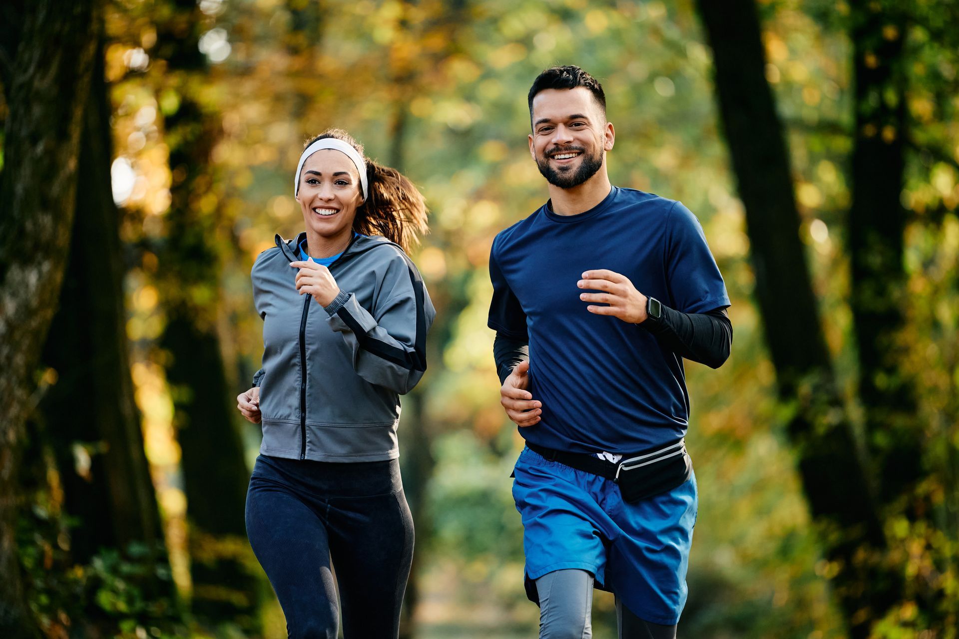 Woman and man jogging smiling in a park with trees.