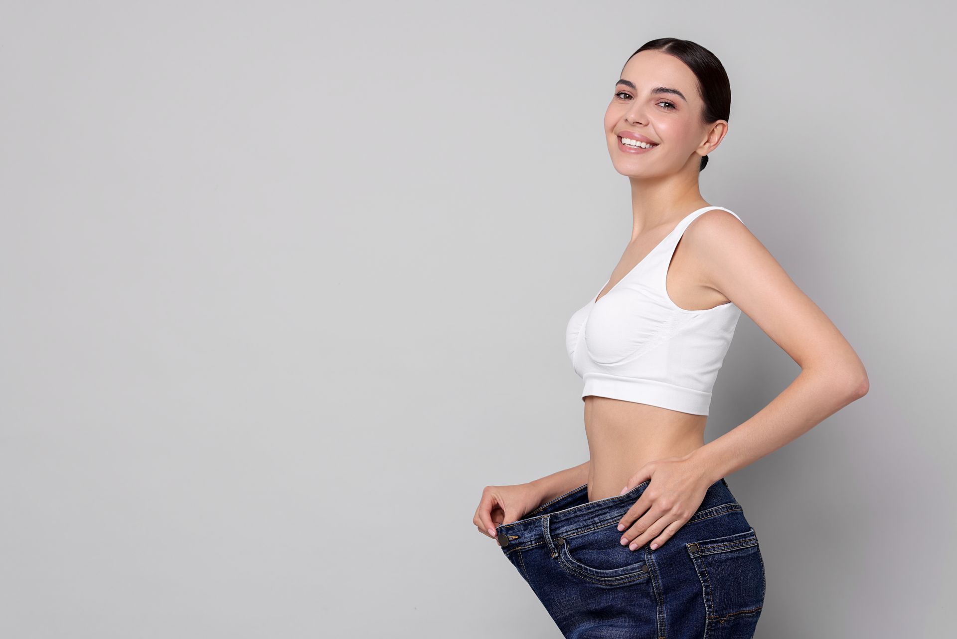 Woman smiling, wearing loose jeans, white crop top; gray background, holding pants showing weight loss.