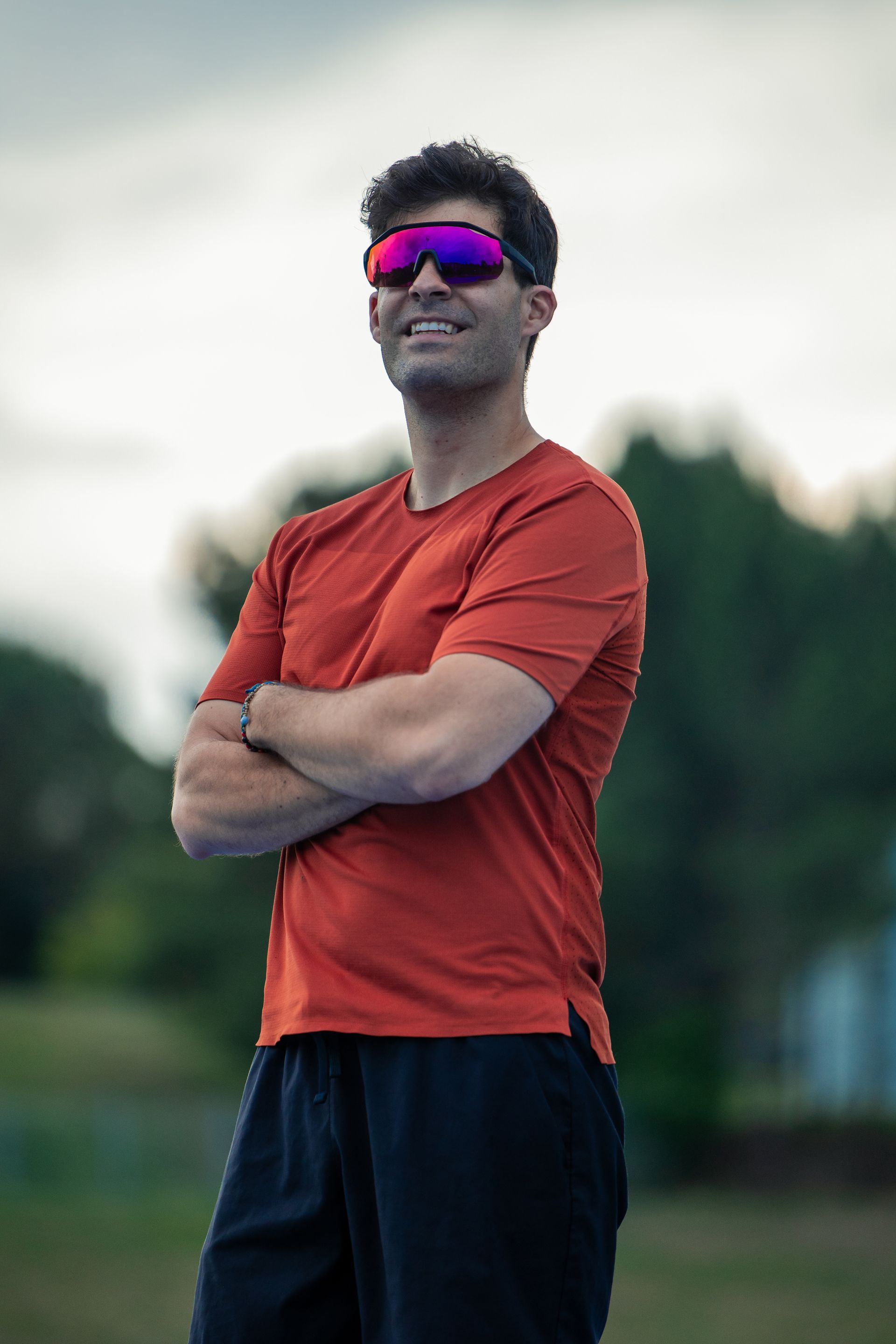 A man wearing sunglasses and an orange shirt is standing with his arms crossed.