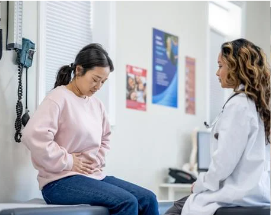 A woman is sitting on a table holding her stomach while talking to a doctor.