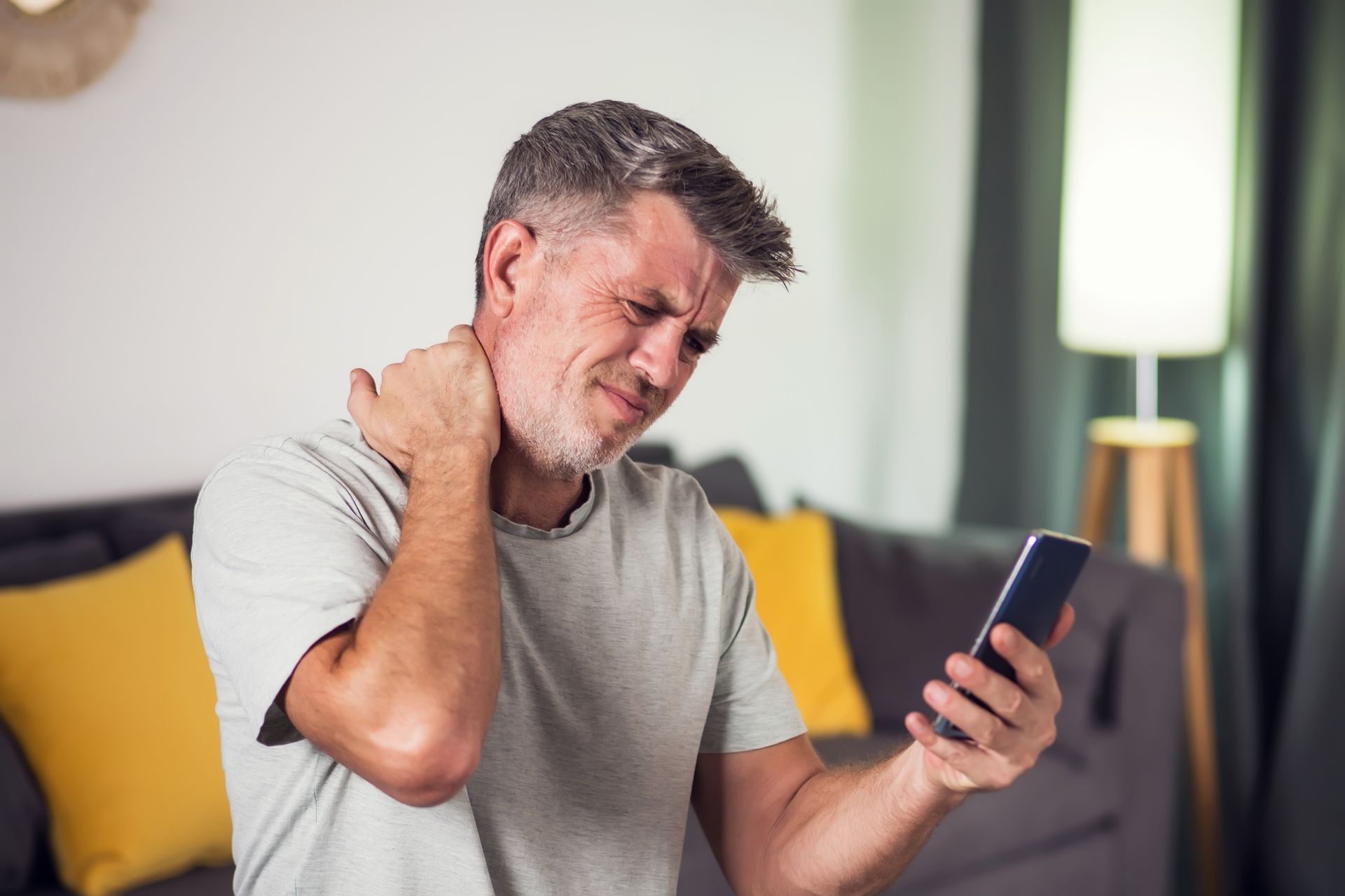 Man on couch grimacing and rubbing his neck while looking at a smartphone