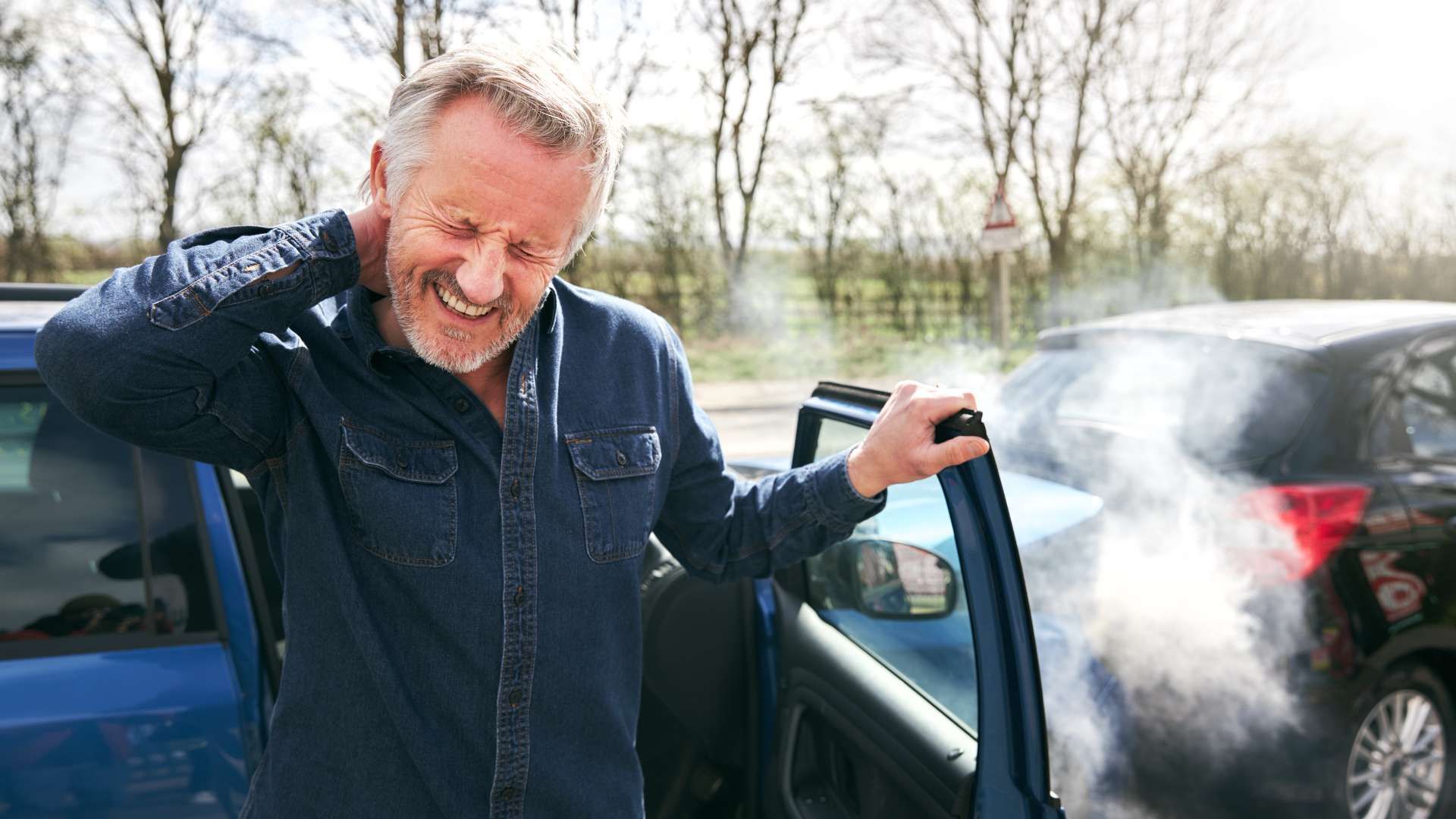 Man smiling beside an open car door with steam or smoke billowing from the vehicle on a cold day