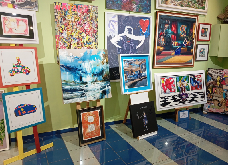 Opere d'arte esposte in una galleria d'arte, tra cui dipinti su cavalletti e appesi alle pareti. Stili colorati e vari.