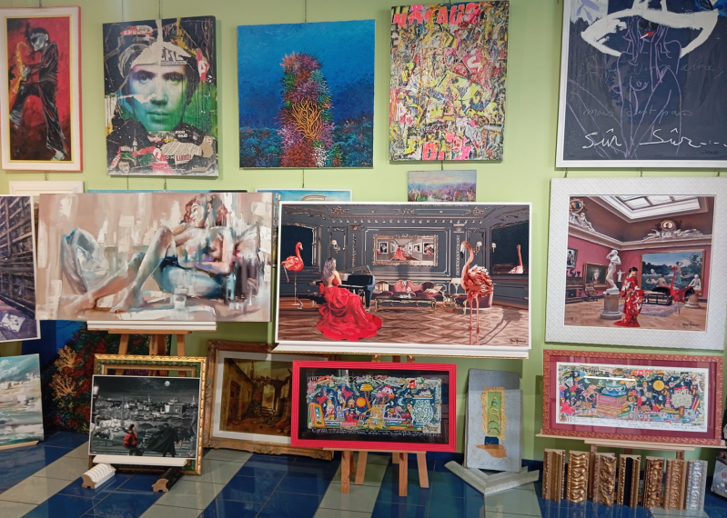 Galleria d'arte con dipinti colorati alle pareti e su cavalletti, tra cui ritratti e paesaggi.