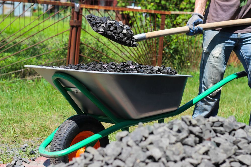 Person Shovels Black Gravel Into a Wheelbarrow on a Sunny Lawn — B&T Earthworks & Civil in Smithfield, QLD
