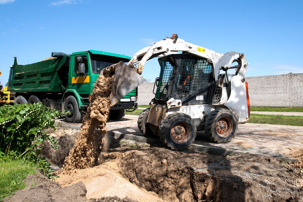 a White Skid Steer Loading Brown Dirt Into a Green Dump Truck in a Field — B&T Earthworks & Civil in Yorkeys Knob, QLD