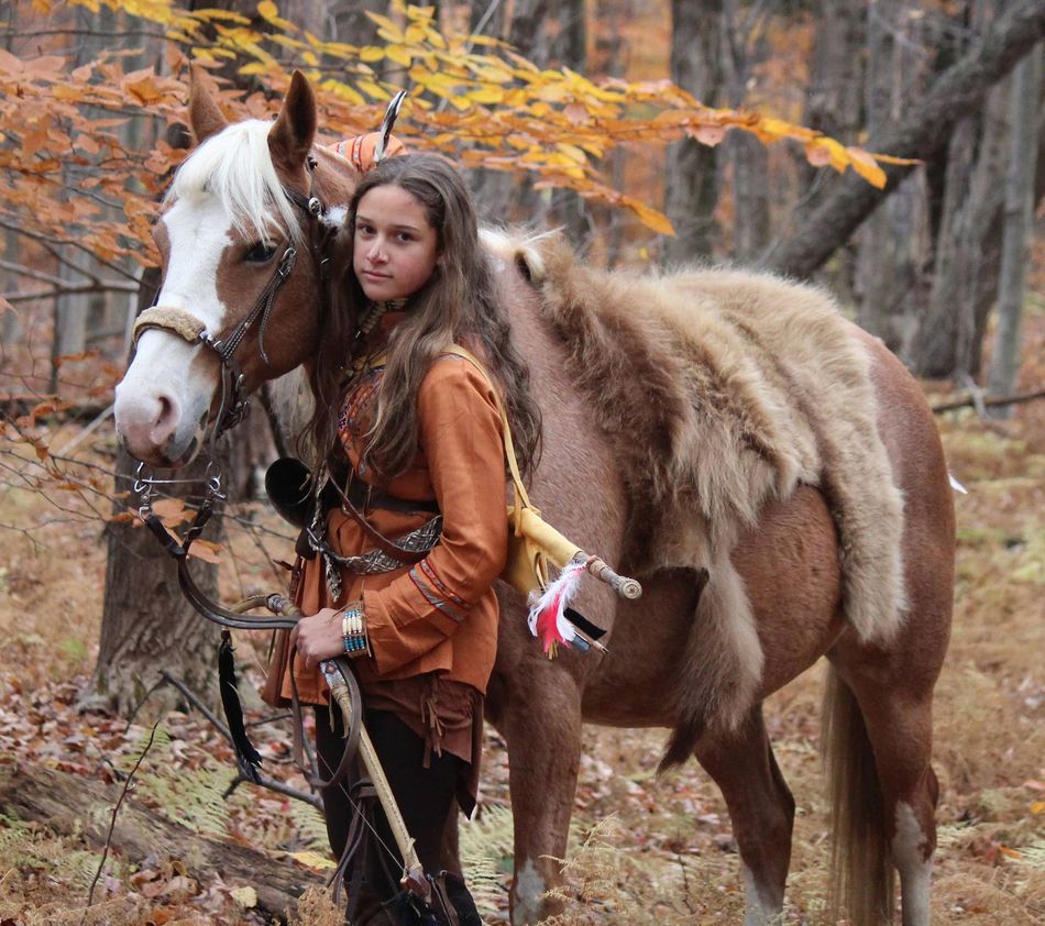 A girl is standing next to a brown horse in the woods