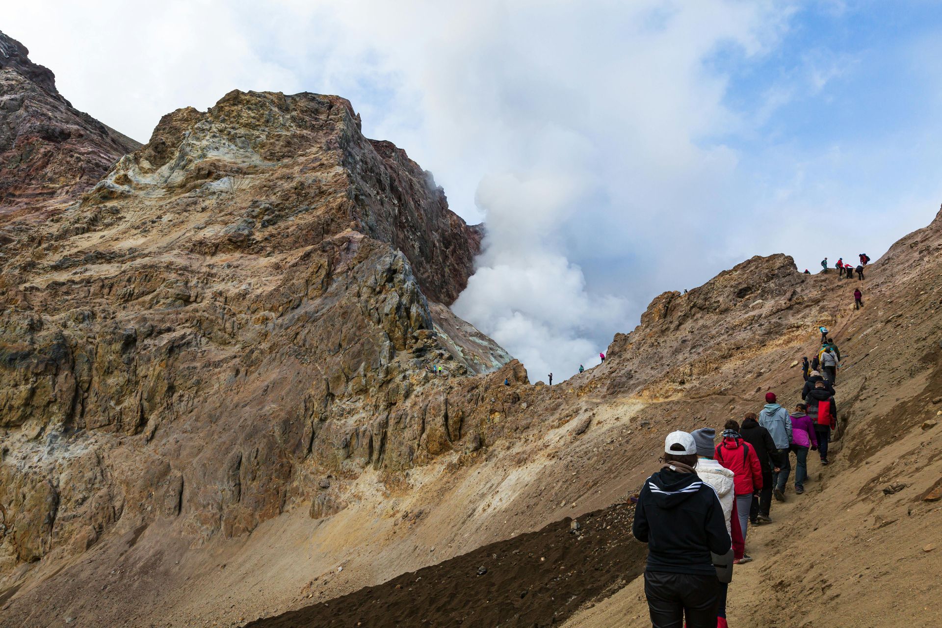 Excursionistas ascienden por un sendero de montaña hacia una zona volcánica humeante bajo un cielo parcialmente nublado.
