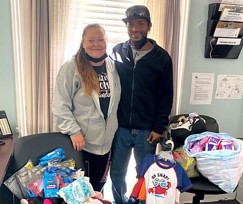 Couple poses with donated baby clothes and supplies in an office setting.