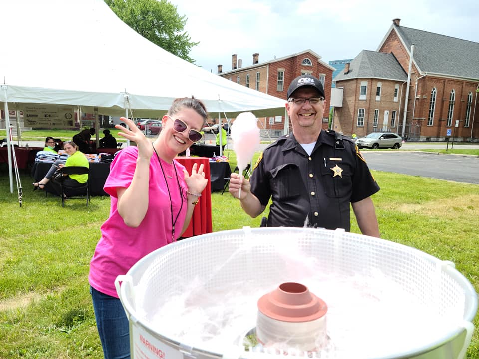 Woman in pink waves, officer holds cotton candy at outdoor event.