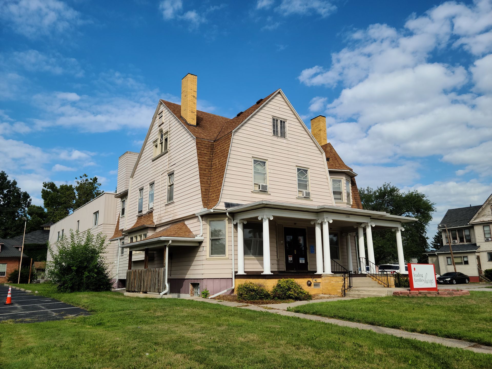Large, light-colored house with a covered porch and a brown roof, set on a green lawn with a blue sky in the background.
