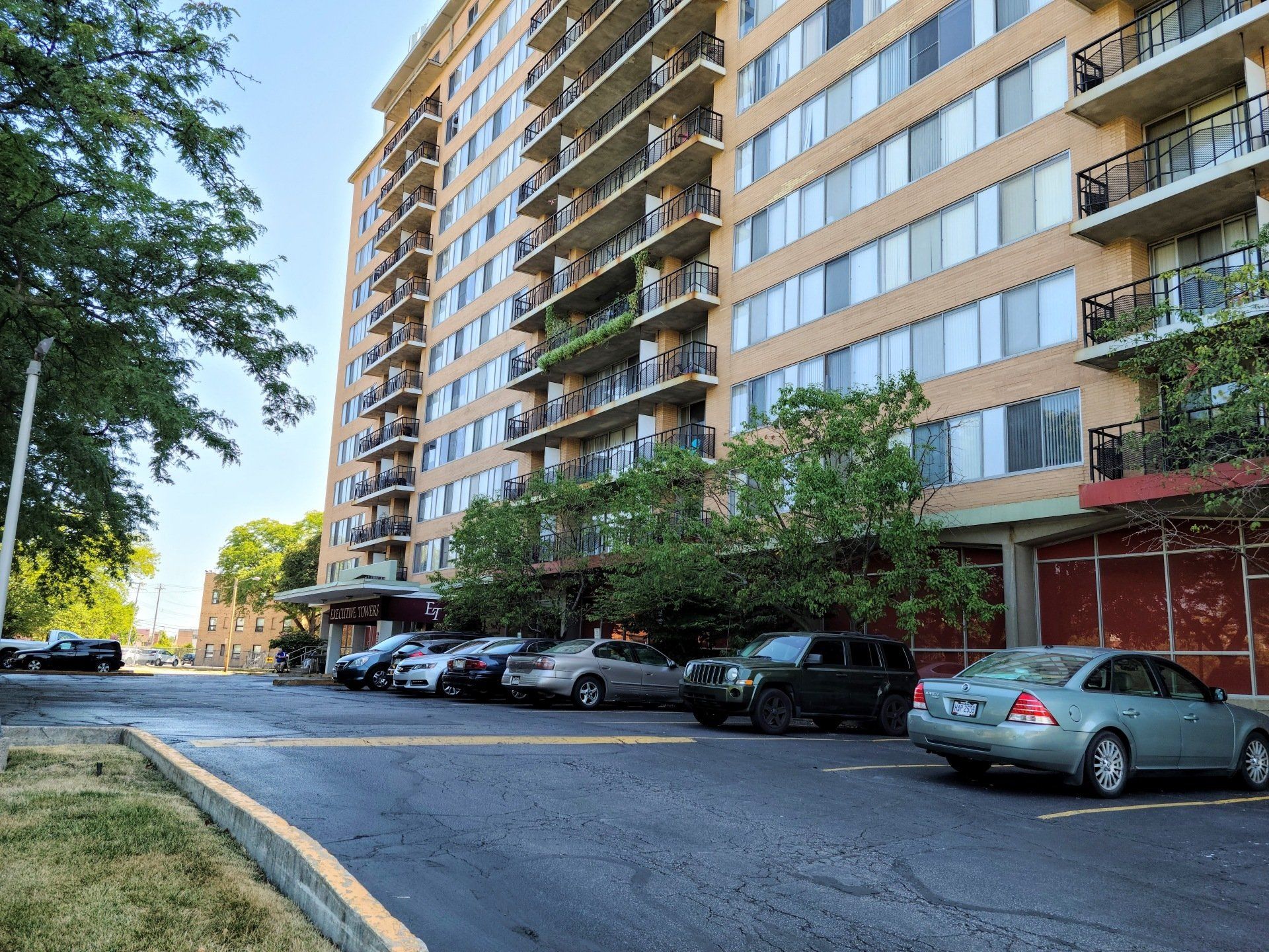 A tall apartment building with balconies, parked cars, and trees. Blue sky visible.