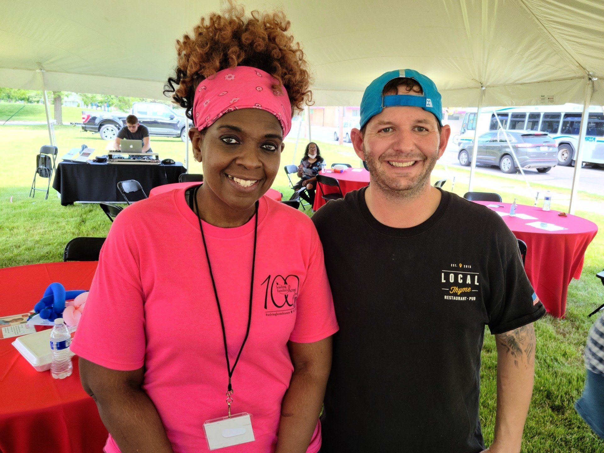 Two people smiling under a tent: woman in pink, man in black. Outdoor setting, daytime.