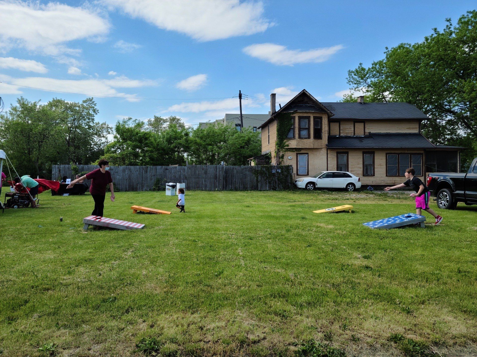 People playing cornhole on a grassy lawn, with a house and trees in the background. Sunny day.