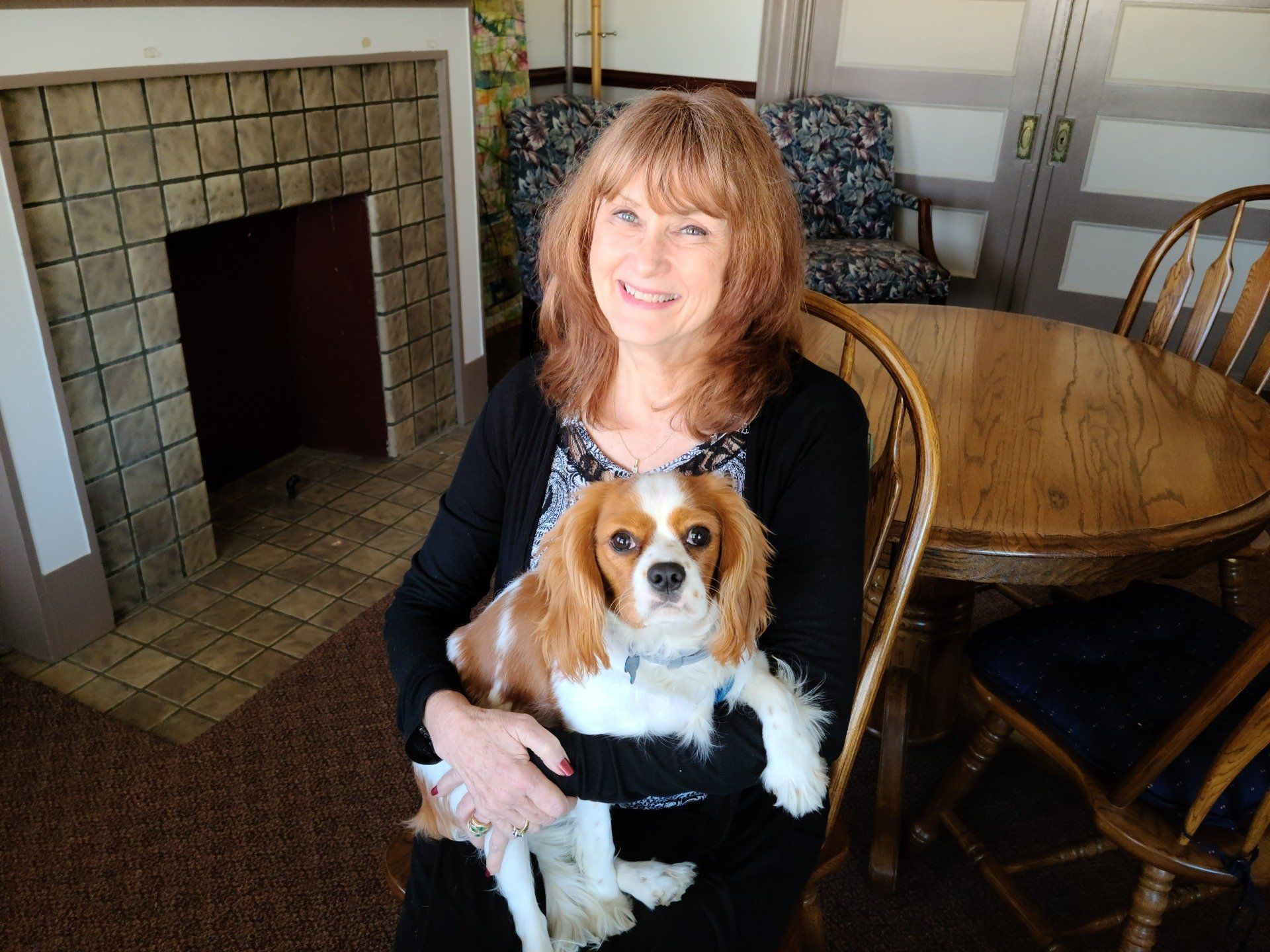Woman with long red hair holding a Cavalier King Charles Spaniel; sitting near a fireplace and wooden table.