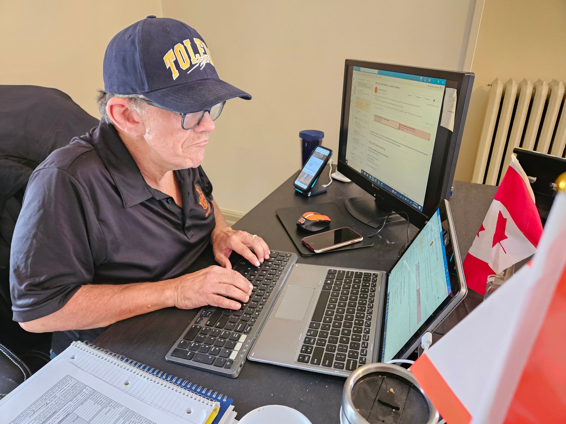 Man in cap, glasses, typing at desk with computer, laptop, Canadian flags.