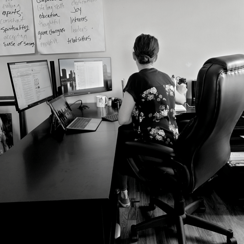 Woman at desk working on computer, two monitors, notes on wall.