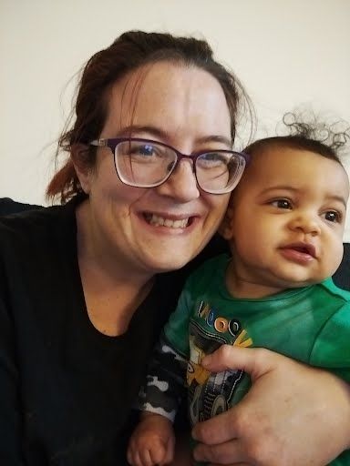 Woman with glasses smiles, holding a baby in a green frog shirt.