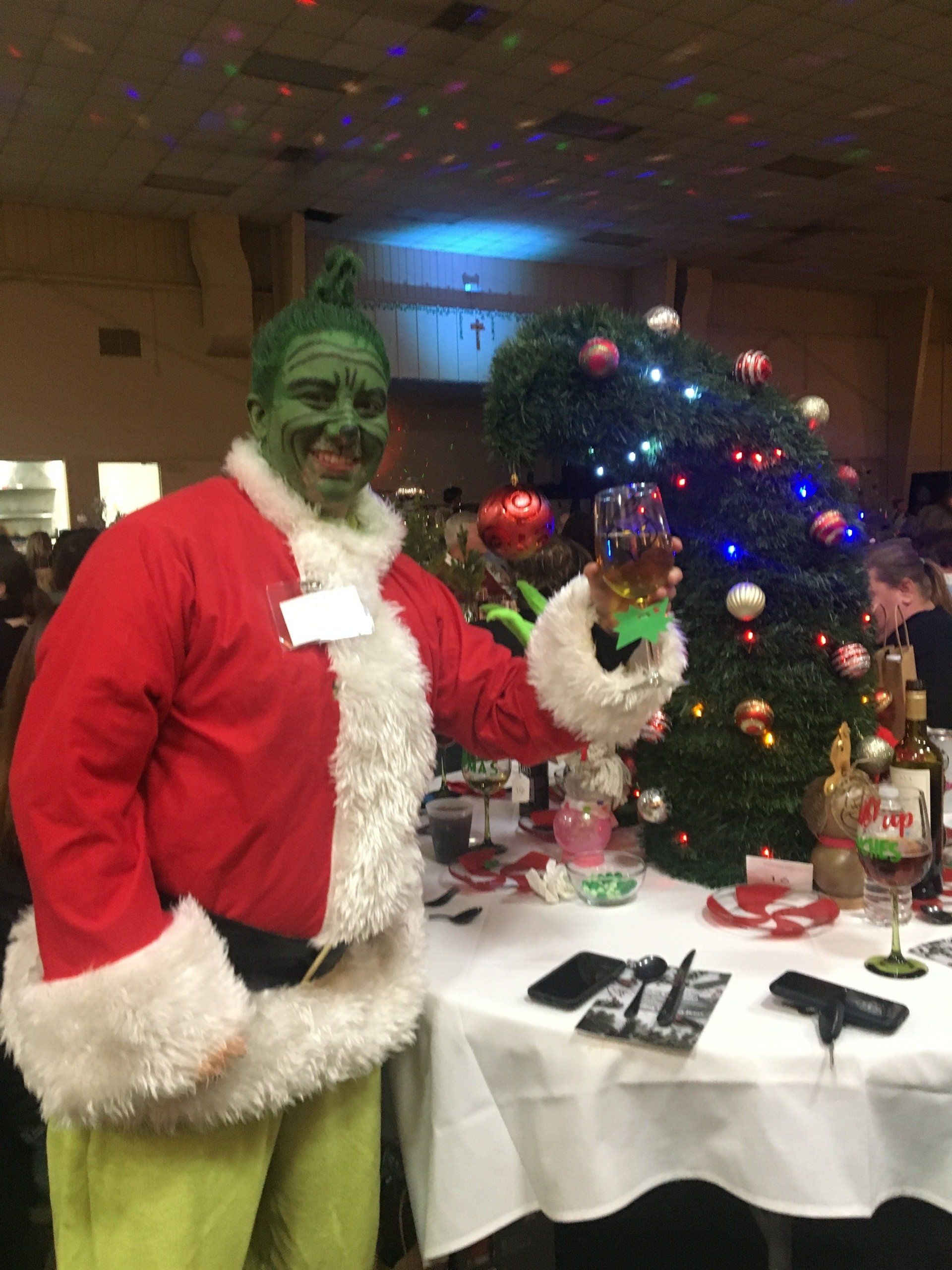 Grinch in Santa suit holds a drink. Green face, red coat, and festive table with Christmas tree in a decorated room.