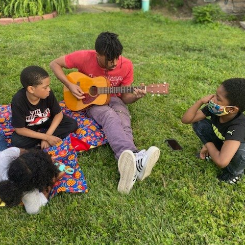 Four children and a teen on a blanket in grass; teen plays guitar, kids watch and listen.