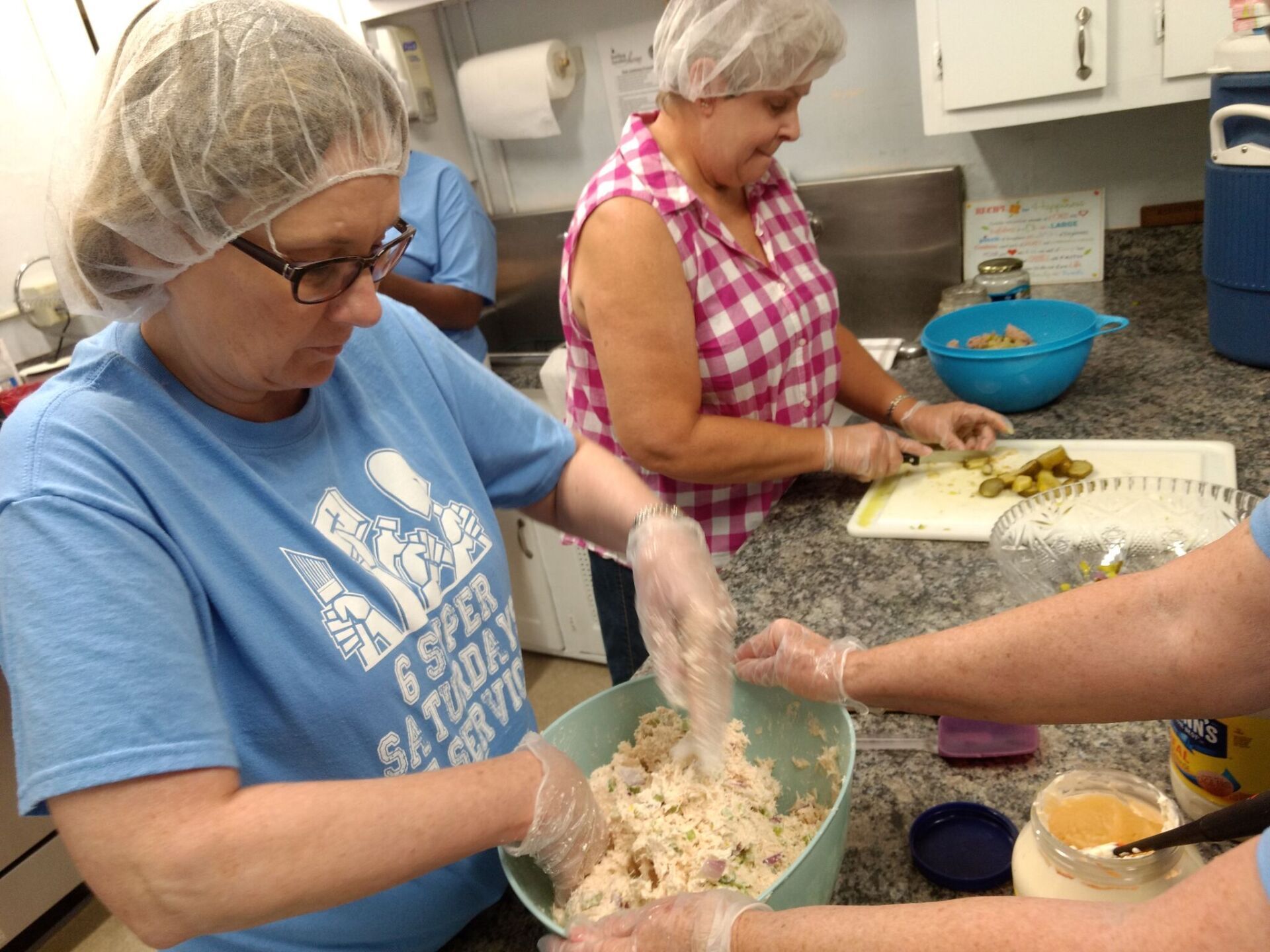 Volunteers in hairnets and gloves prepare food in a kitchen. Woman mixes ingredients, another slices pickles.