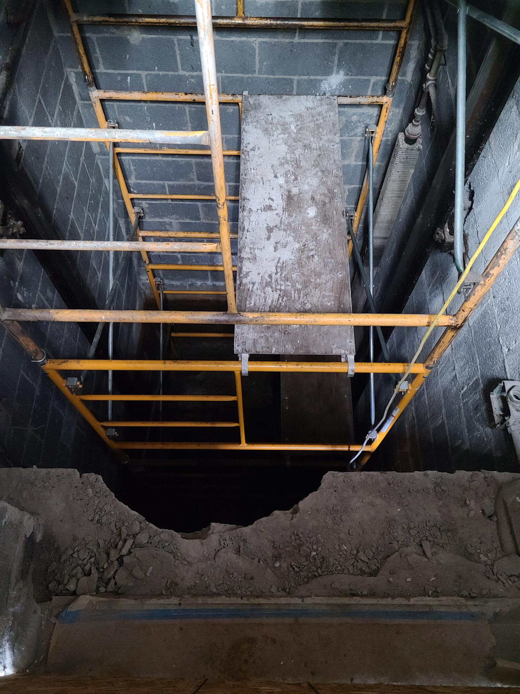 Yellow scaffolding frames a dark, deep pit, with a ladder, against a backdrop of gray walls.