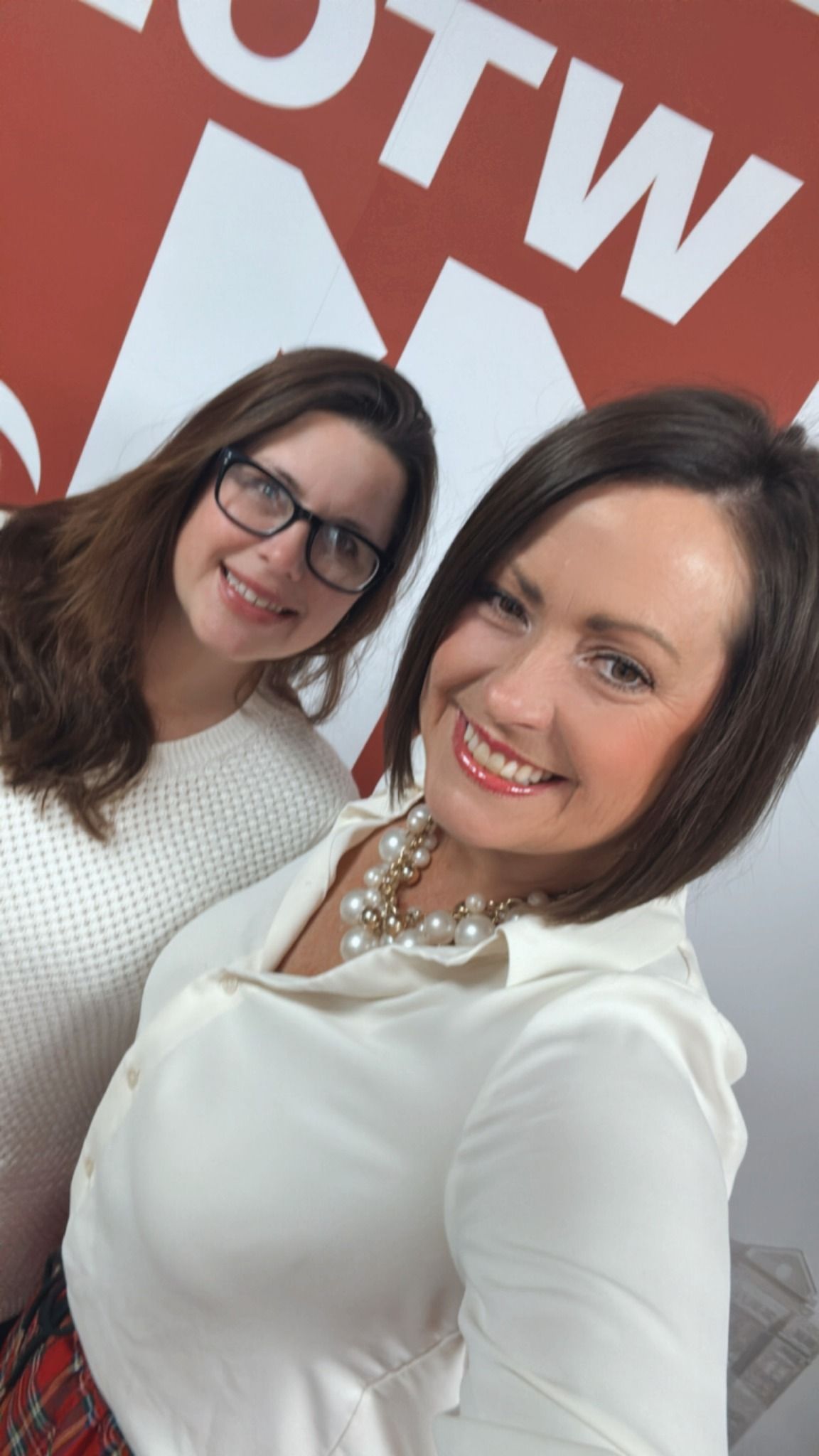 Two women smiling; one wearing glasses and white sweater, the other with short hair, white blouse, pearl necklace. Red and white background.