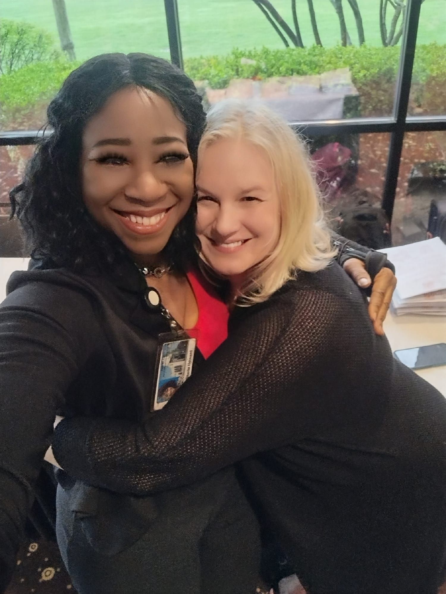 Two women, one Black, one white, embrace and smile. Inside, near a window.