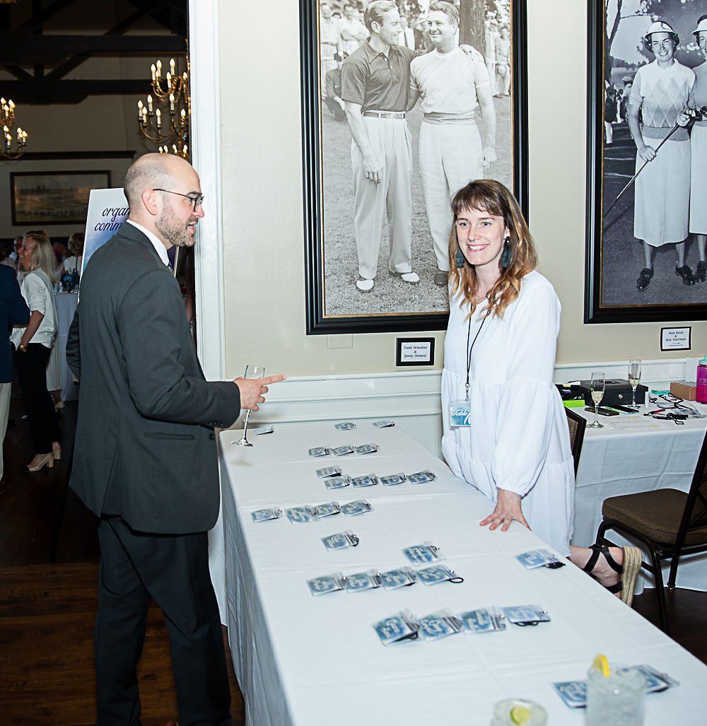 Man in suit gestures toward a table with a woman smiling. They are at an event with photos on the wall.