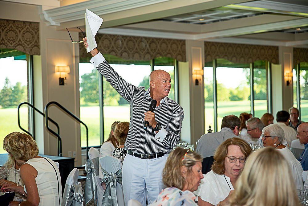 Man in striped shirt holds paper and microphone, speaking to a crowd in a well-lit dining room.