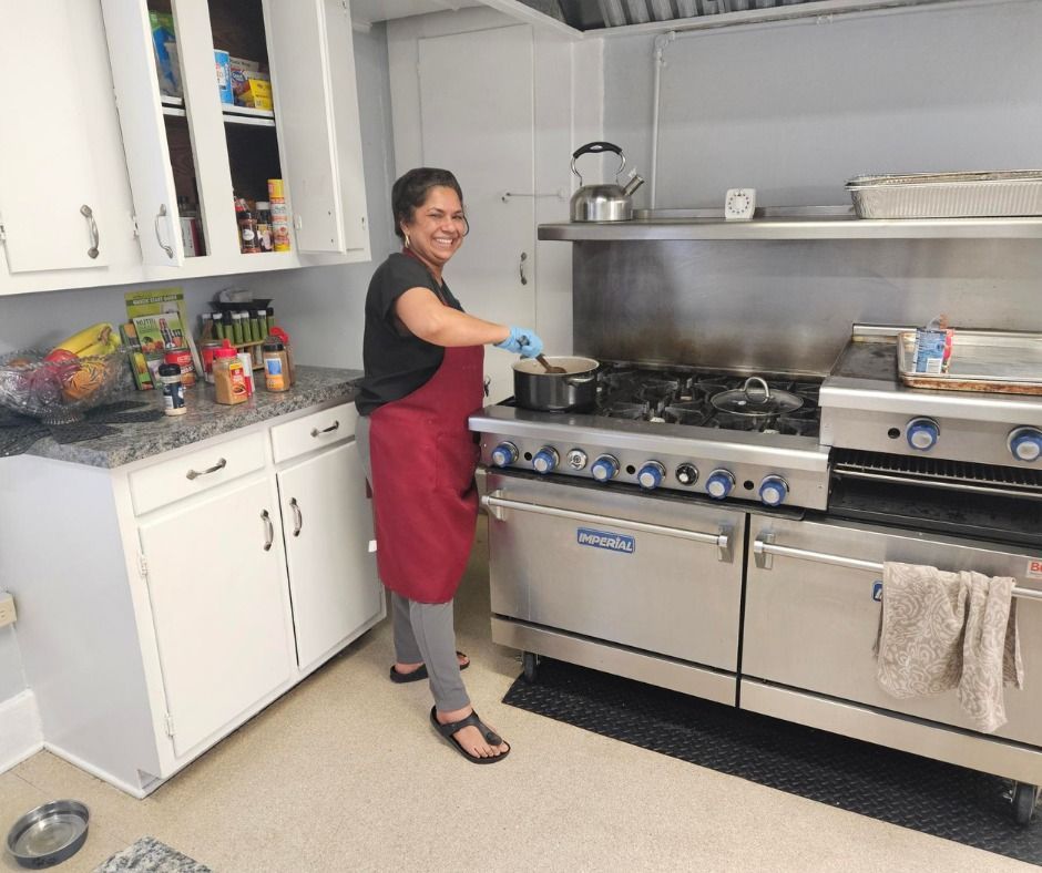 Woman in a kitchen, wearing an apron, cooking on a stove. She is smiling.