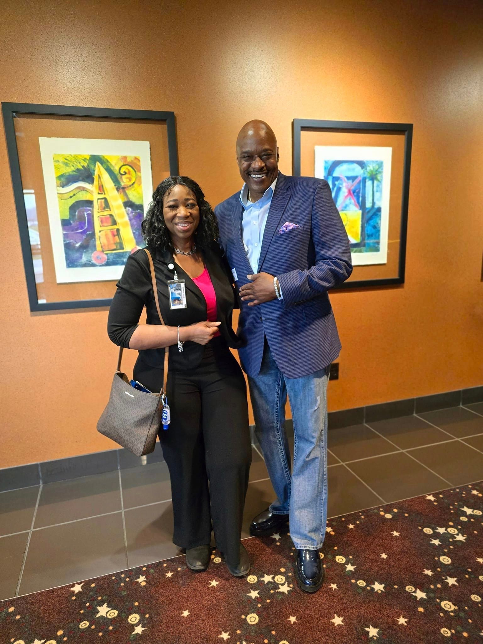 A woman and a man pose in front of framed artwork, smiling. They are in a room with patterned carpet.