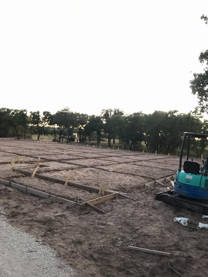 Construction site: concrete forms set in dirt, small excavator, trees and overcast sky in background.
