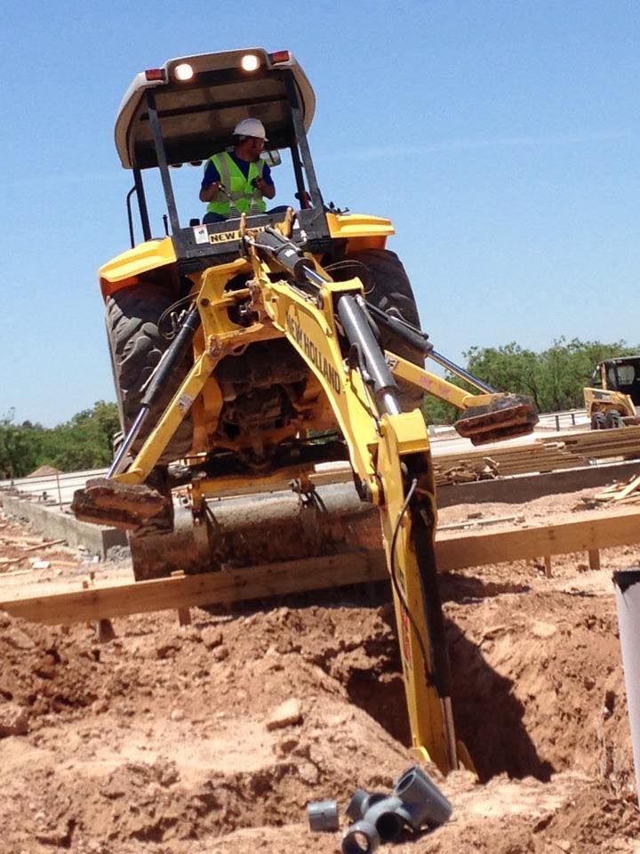 A backhoe digging a trench on a construction site. The operator is wearing a vest and helmet.