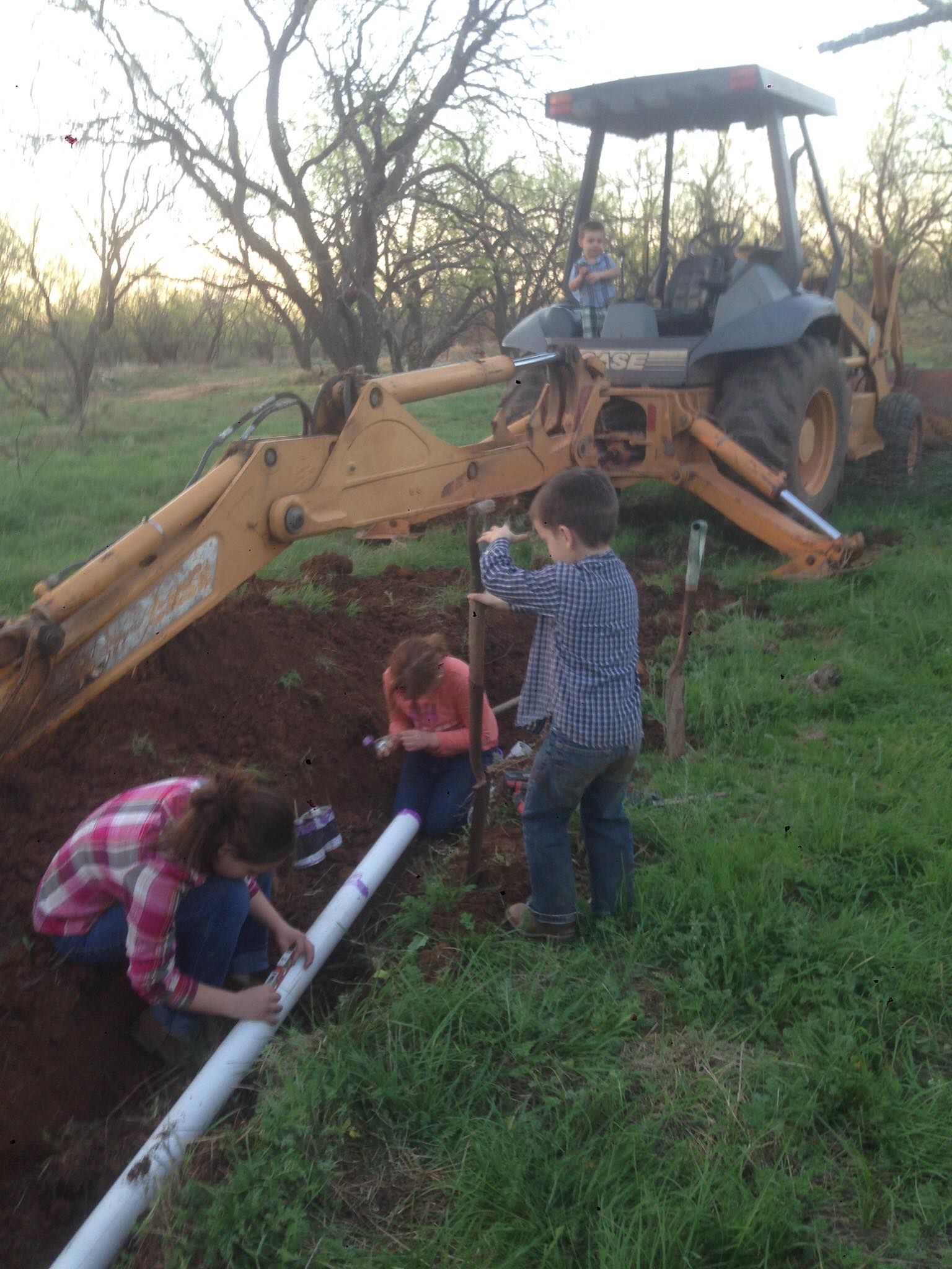 Children work on a pipe in a trench, as a backhoe sits nearby.