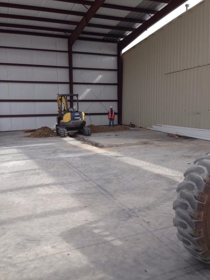 Interior of a metal building with an excavator digging; a worker stands nearby.