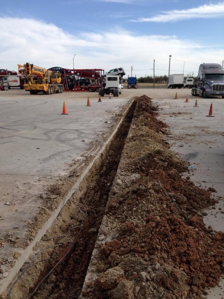 Trench dug in a parking lot, dirt piled beside it. Construction cones and trucks in the background.