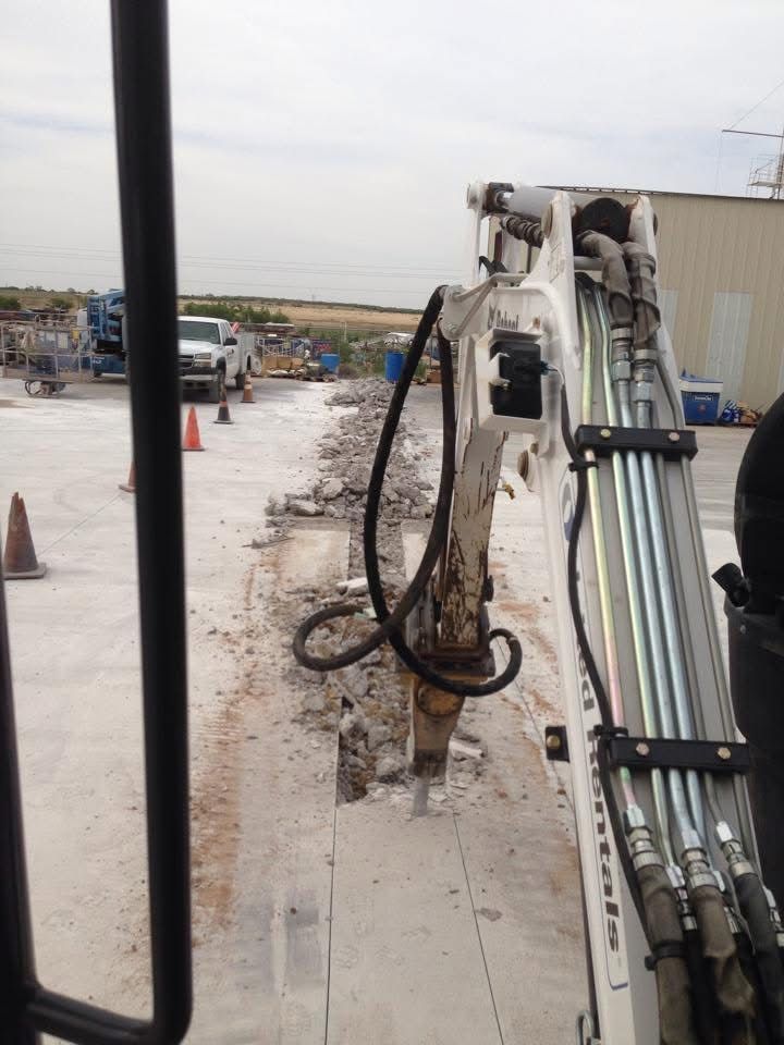 A backhoe breaking concrete on a paved surface with a building and debris in the background.