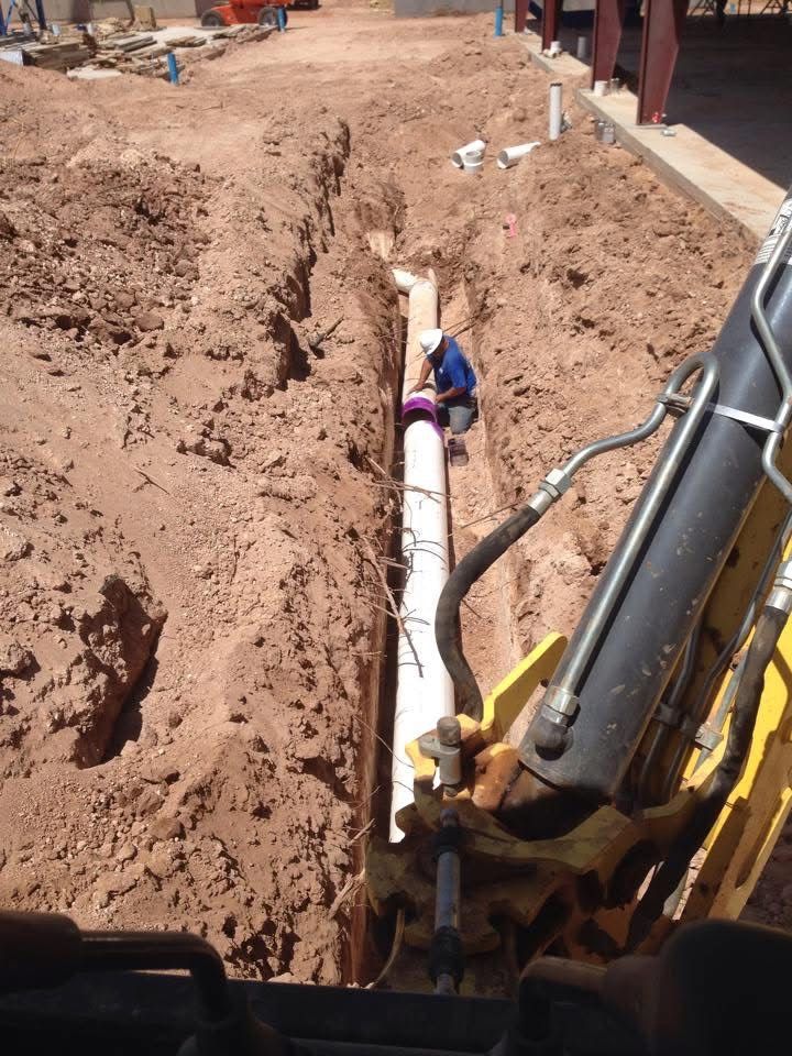 Construction worker installing pipe in a trench, using a backhoe in a dirt setting.