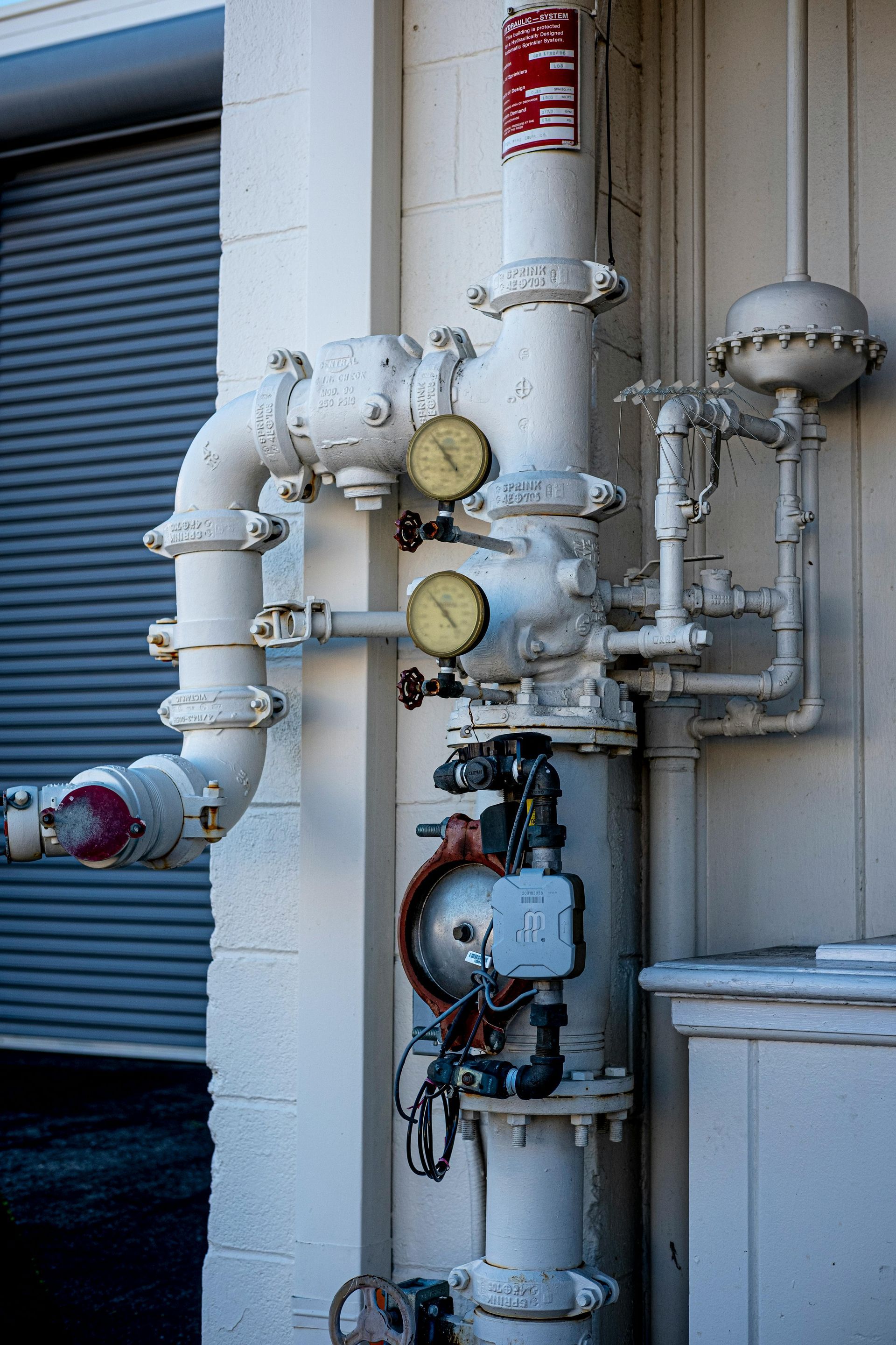 White painted industrial piping system with gauges and a vertical stack, mounted outdoors on a wall.