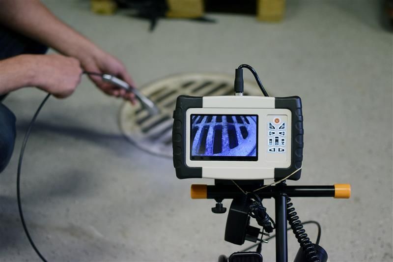 A person using an inspection camera to look at a drain, displayed on a screen mounted on a tripod.