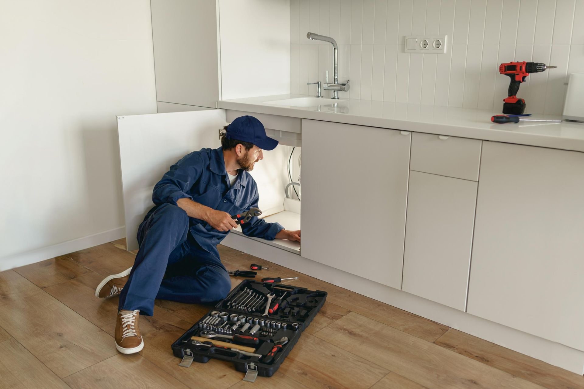 A plumber in blue coveralls works under a kitchen sink. Tools and drill lay nearby.
