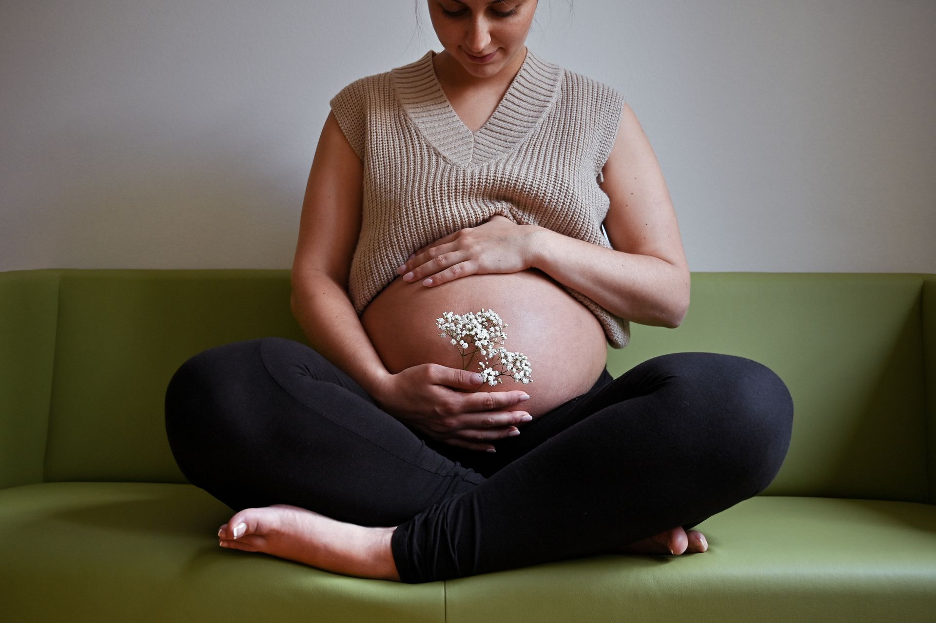 Schwangere Frau sitzt im Schneidersitz auf einem grünen Sofa, die Hände umschließen ihren Bauch und halten kleine weiße Blumen. Kirstin Hammerstein, hammerstein-pictures