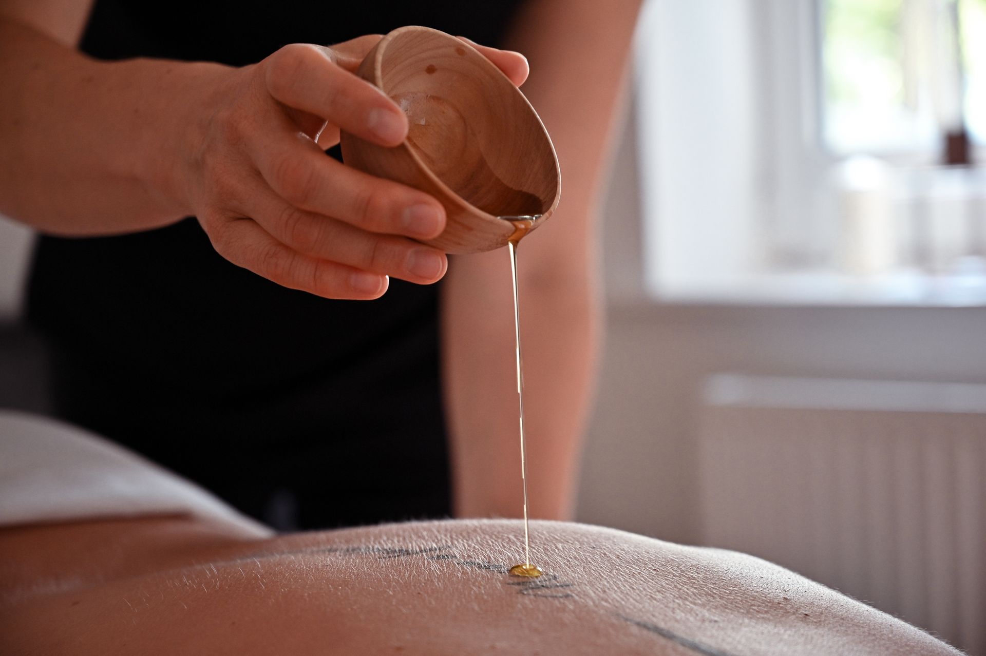 Person, die während einer Massage Öl aus einer Holzschale auf den Rücken einer anderen Person gießt.
