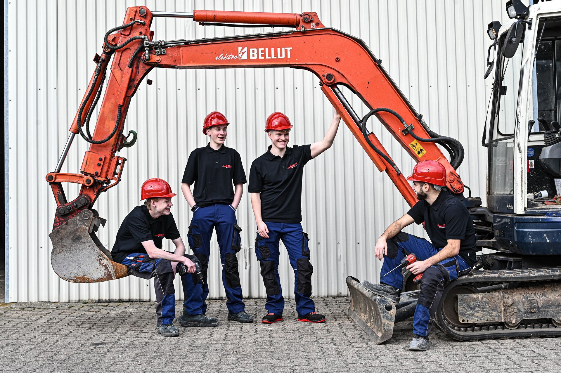 Vier Bauarbeiter posieren neben einem roten Bagger, alle tragen Helme und Arbeitskleidung. Kirstin Hammerstein, hammerstein-pictures