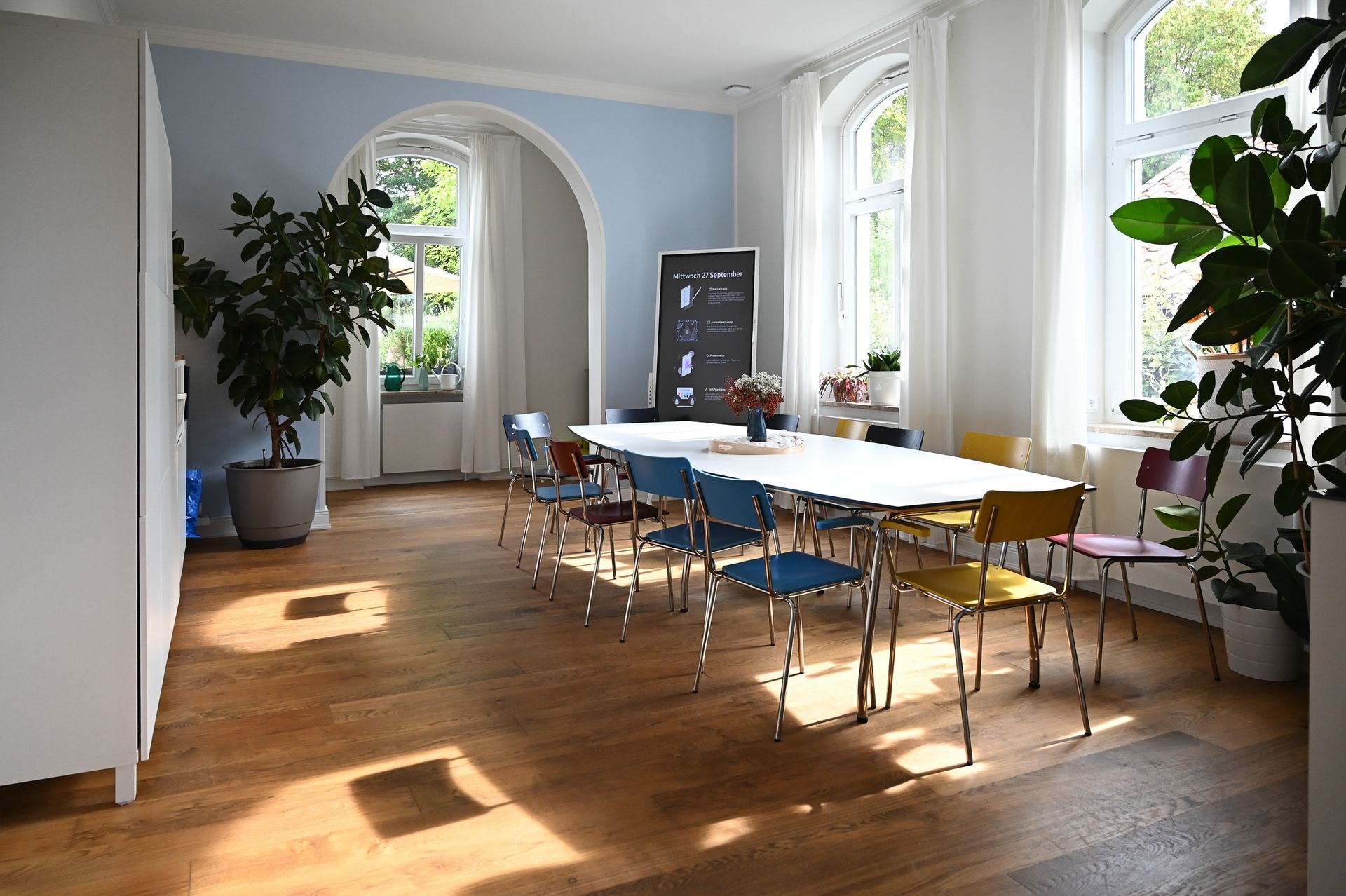 Heller Raum mit langem Tisch, bunten Stühlen, großen Fenstern, Pflanzen und einer Tafel. Kirstin Hammerstein, hammerstein-pictures