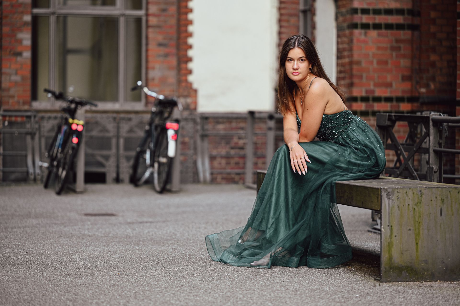 Frau im smaragdgrünen Kleid sitzt auf Betonbank neben Fahrrädern, im Hintergrund ein Backsteingebäude. Kirstin Hammerstein, hammerstein-pictures